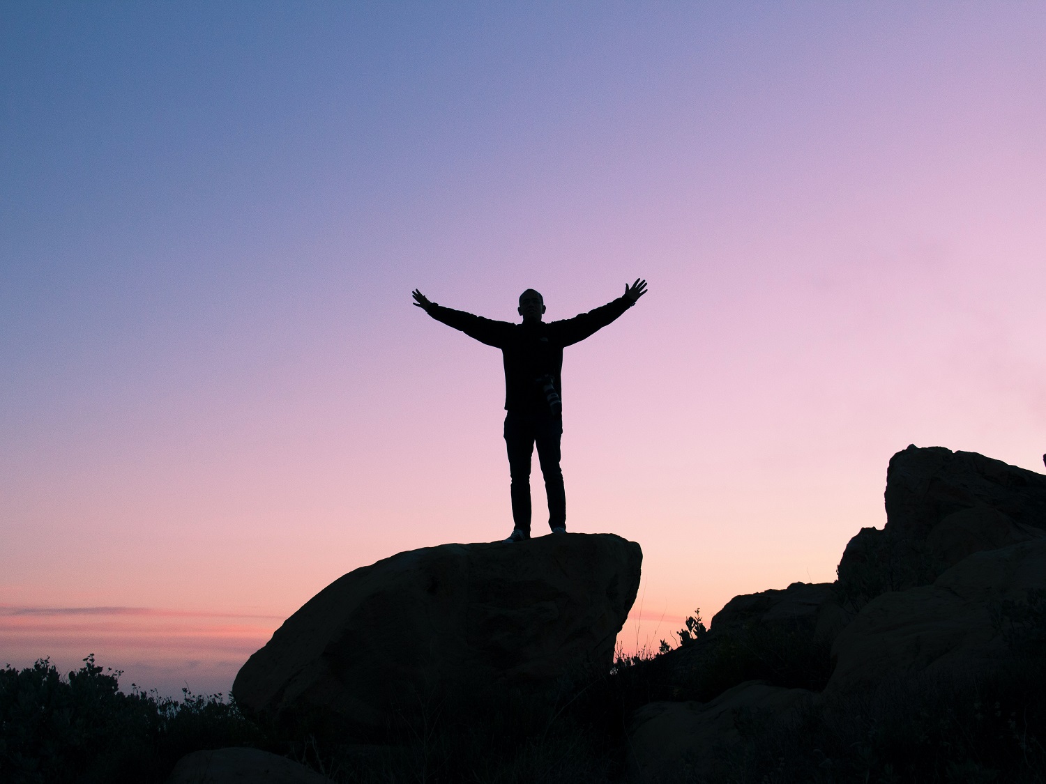 Person standing triumphantly on a rock with arms raised against a dramatic golden hour sky
